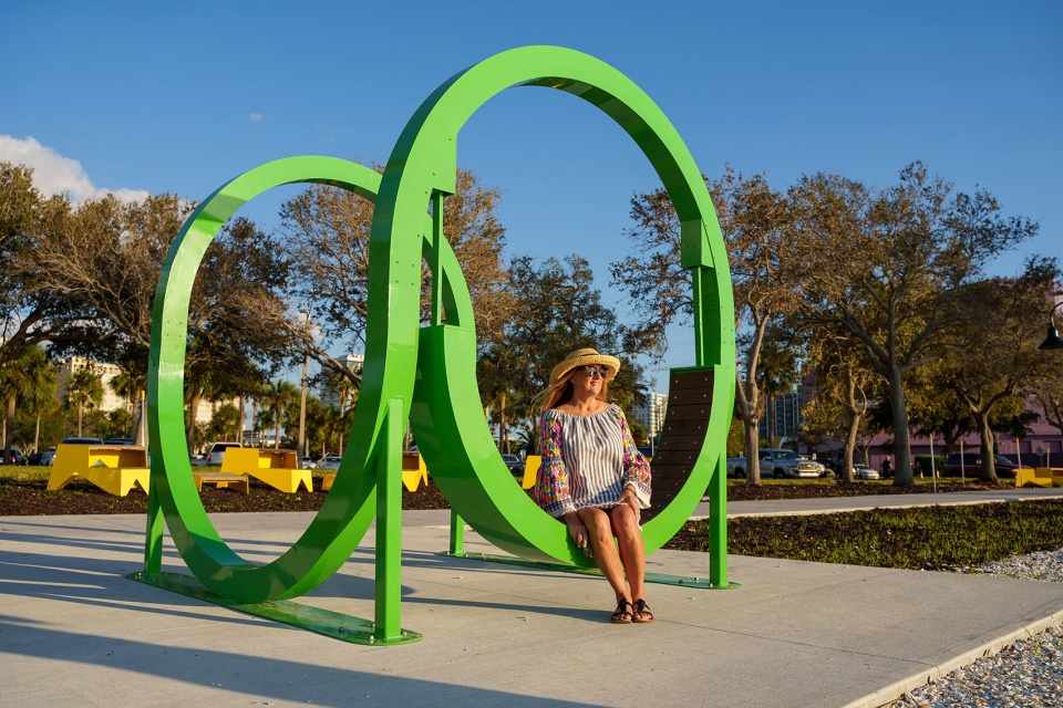 Interlocking green steel ring seats in a waterfront park with a visitor sitting in one, playable art by The Urban Conga